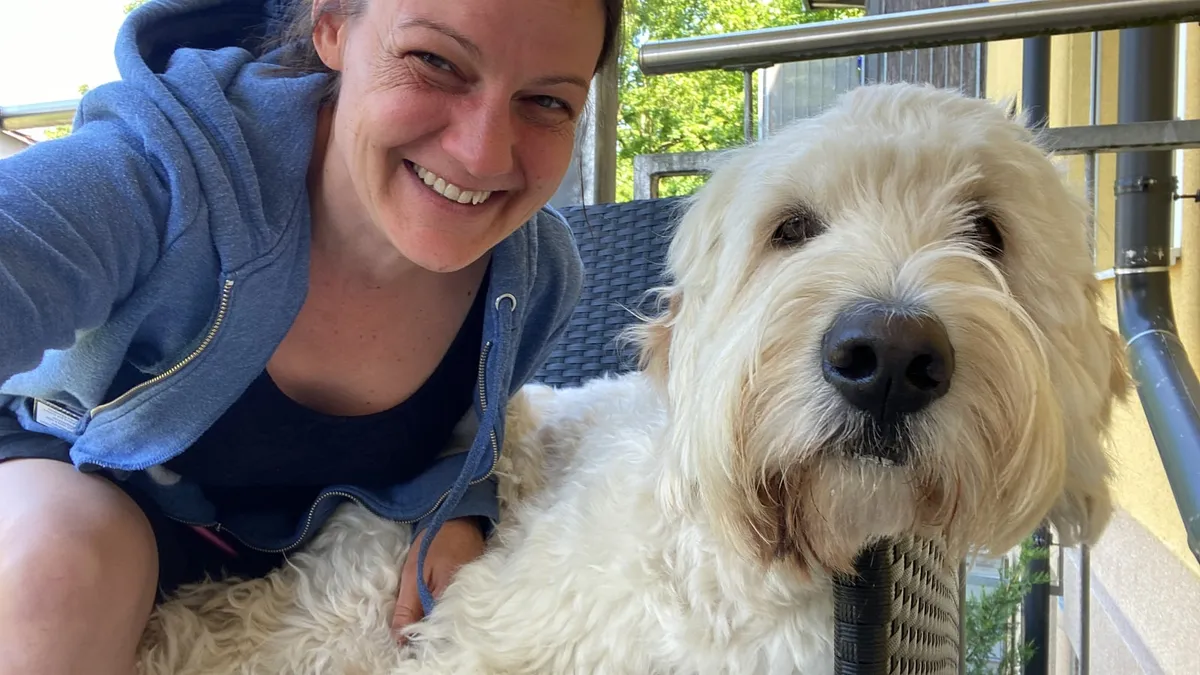 Eva lacht in die Kamera, neben ihr ein heller, gelockter Hund, der den Kopf auf eine Stuhllehne legt.